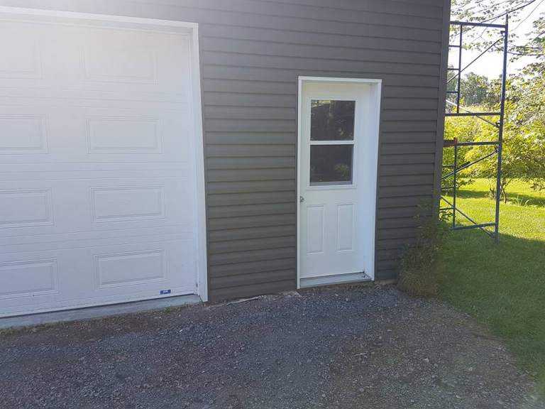 Close-up of a house corner freshly fitted with dark taupe vertical vinyl siding, a white garage door, a new white side door, and a scaffold tower alongside