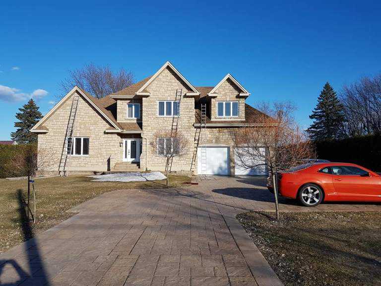 Two-storey stone-clad house in late winter with extension ladders set up against the roof and an orange sports car parked in the driveway