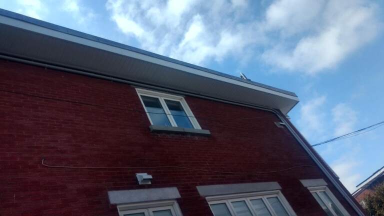 Débord de toit en contre-plongée : soffite ventilé et fascia clairs sur brique rouge — Gouttières et Revêtements Synergy